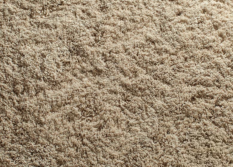 Beiger Teppich mit hoher, flauschiger Oberfläche und unregelmäßiger, lebendiger Struktur.